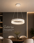 A modern circular LED pendant light with a white frame and sparkling clear crystal-like accents, hanging over a dining table in a stylishly decorated room with warm ambient lighting.